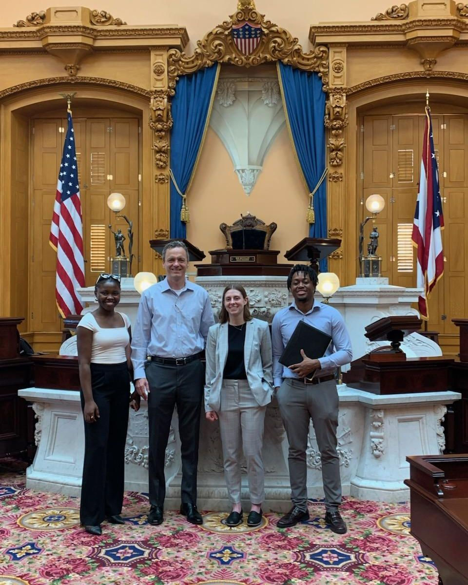 Three people in the Ohio Senate Chambers are shown. OIP Fellow Adesewa Adeyefa stands near Jon Barron, chief of staff and general counsel for the majority caucus in the Ohio Senate, and Caroline Waller and Kofi Agyepong 
