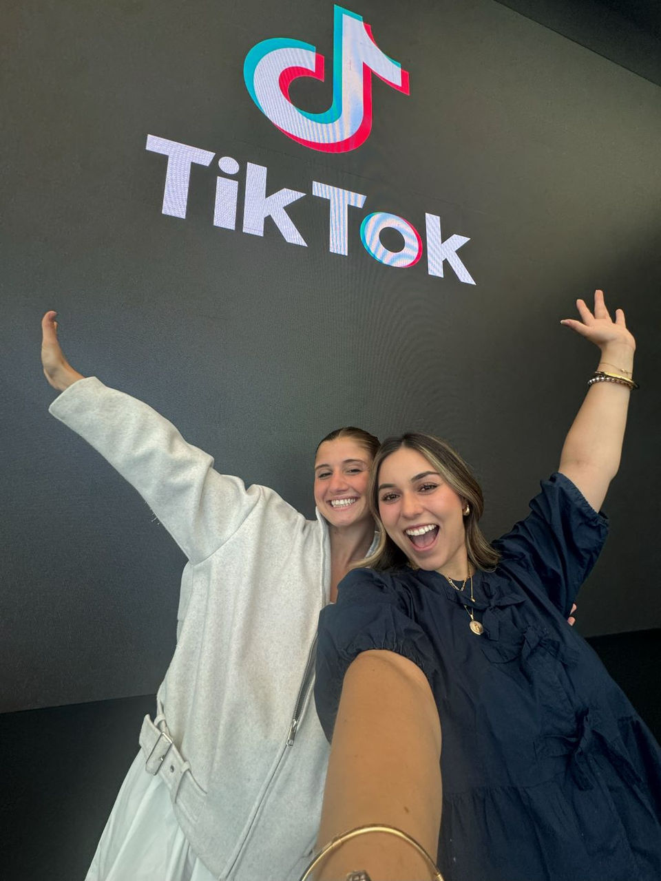 Two college students Delfina Lassalle and Daniela Padilla with their arms outstretched with the TikTok signage behind them.