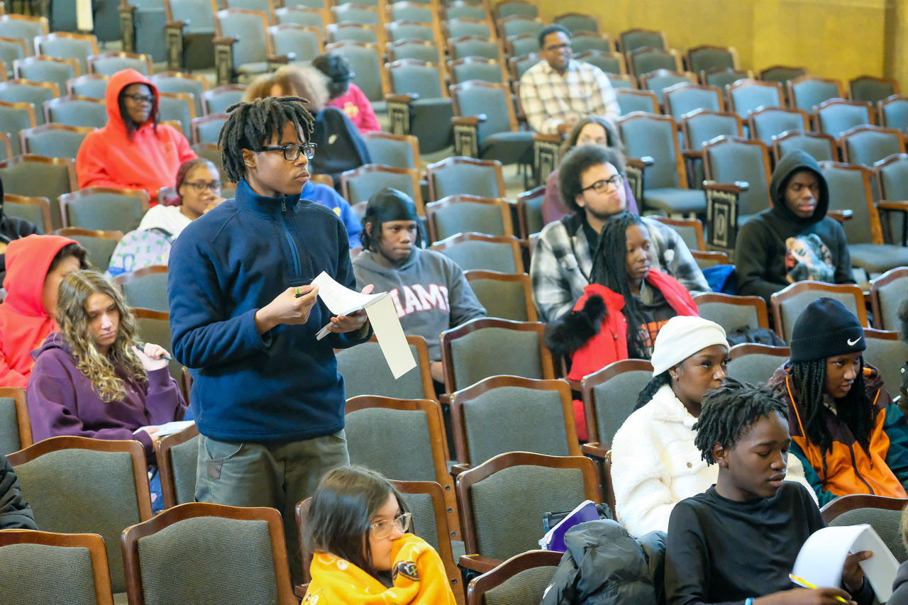 A student holding a reporter's notebook asks a question in an auditorium surrounded by other students.