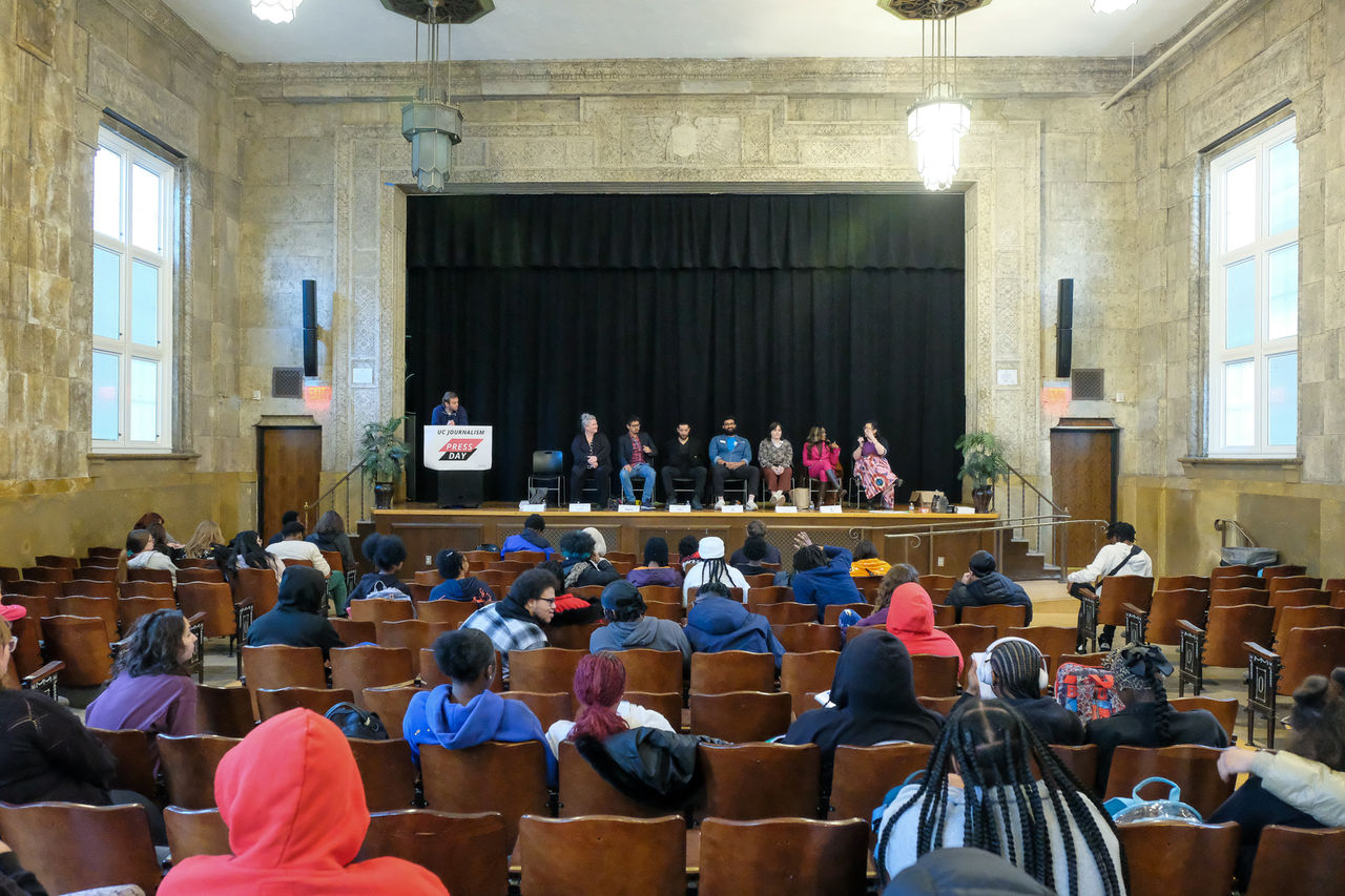A wide shot of the stage shows panelists with members of the audience in an auditorium.
