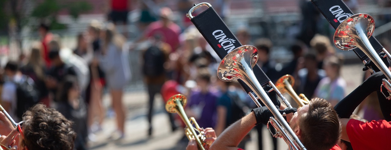 Several UC students and parents are shown standing along UC Main Street while the UC band plays with close ups to trumpets and trombones 