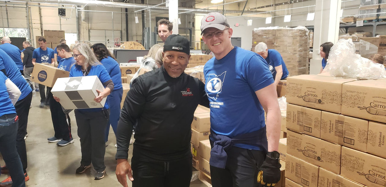 Daniel Cummins is shown smiling with arm around BYU supporter at Fishwick warehouse. There are boxes of food in the background with volunteers stacking items