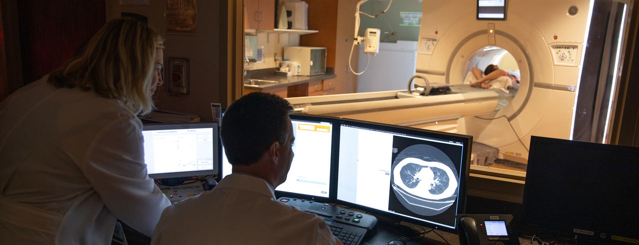 Two doctors review imaging data on a computer while a patient lays in an imaging machine
