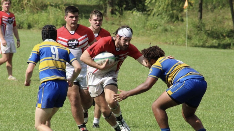 Nursing student Kaleb Kiely running with the rugby ball with two teammates behind him and two opponents trying to tackle him