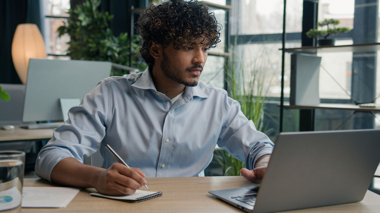 Indian Arabian man accountant writer business entrepreneur businessman write notes in notebook latino guy writing ideas report paperwork on office table studying online with laptop educational course. High quality 4k footage