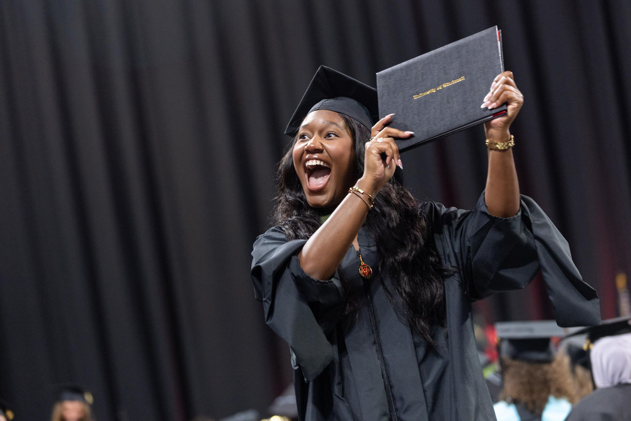 A graduate in a cap and gown holds up her diploma while reacting on stage at Fifth Third Arena.
