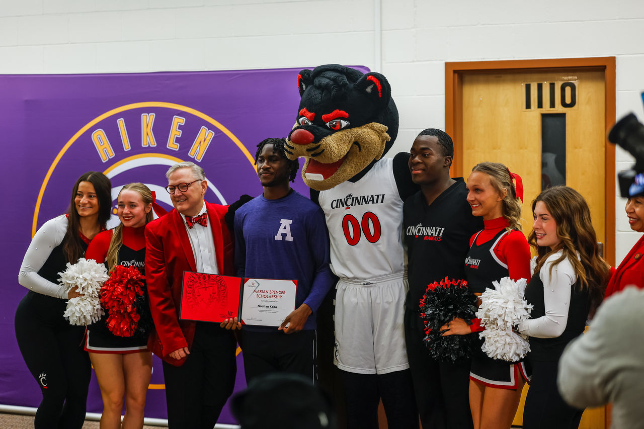 Nouhan Kaba stands holding a Marian Spencer Scholarship award as Jack Miner is standing nearby on one side while the Bearcat Mascot is on the other. Several other UC cheer team members in the photo
