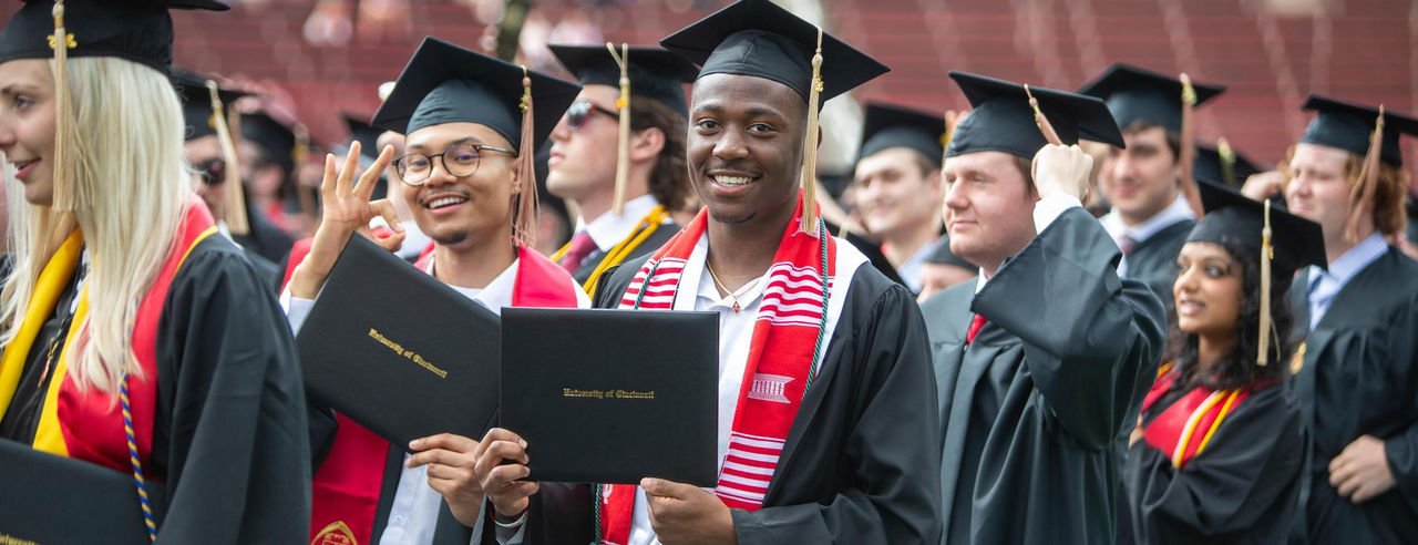 University of Cincinnati graduates smiling and holding diplomas at commencement ceremony.