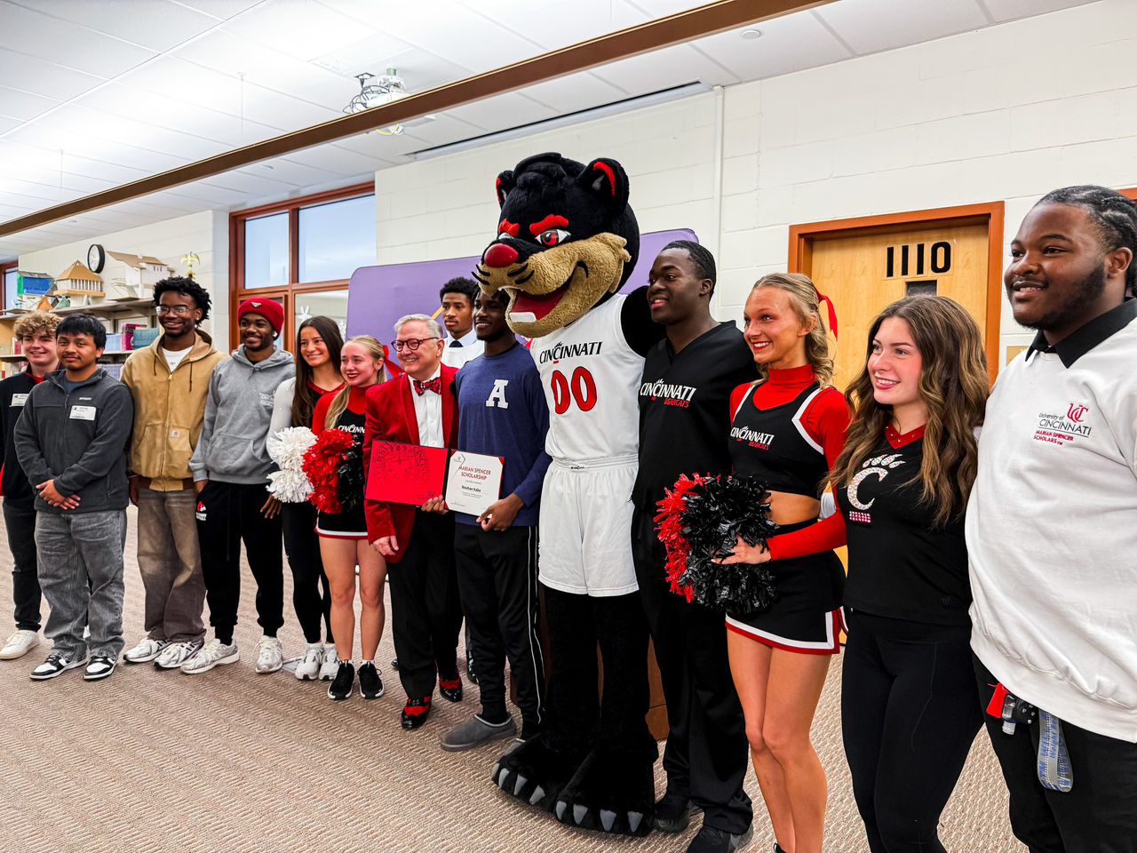A dozen people stand in line for a photo that shows Spencer Scholar Nouhan Kaba, the Bearcat Mascot, UC officials, cheerleaders and Aiken High school fans