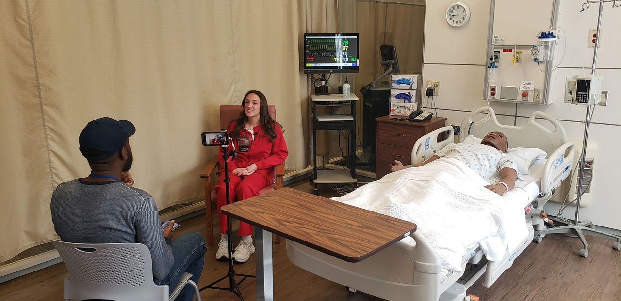image of Javari Burnett sitting with his back to us speaking with Megan Romero, who is seat next to a simulated patient in the College of Nursing simulation lab