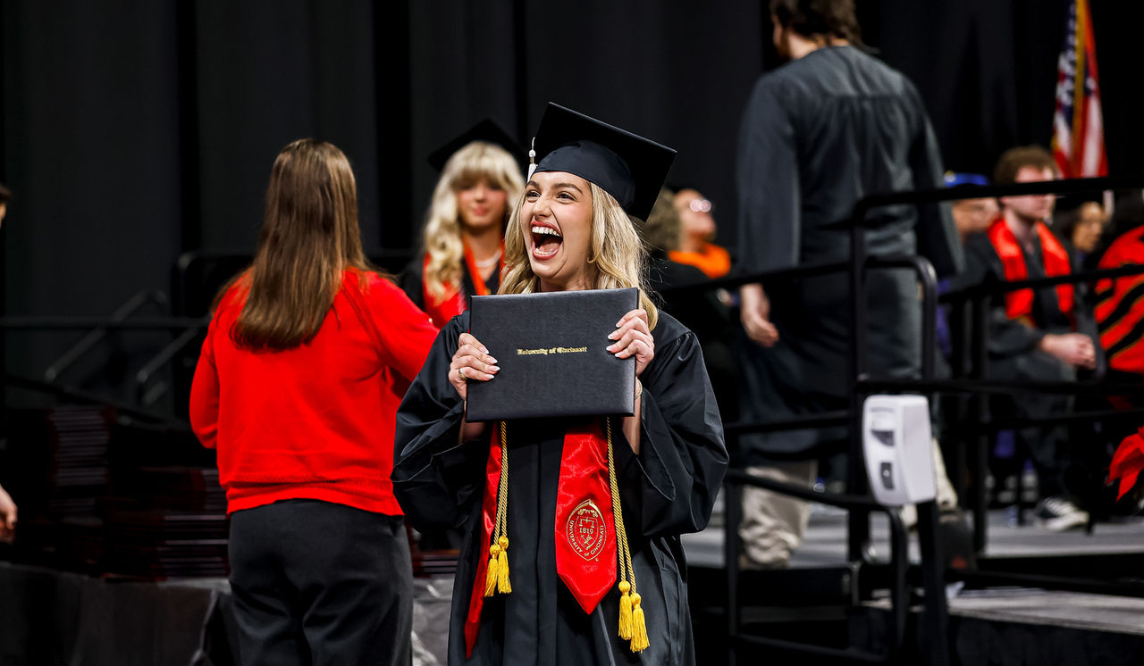 A UC student in cap and gown holds up her diploma in triumph on stage at Fifth Third Arena.
