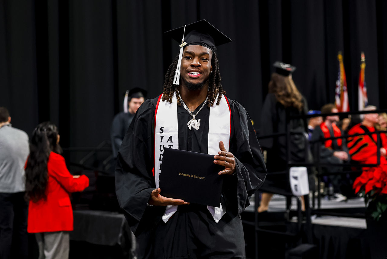 A UC student wearing a student athlete sash holds up his diploma.