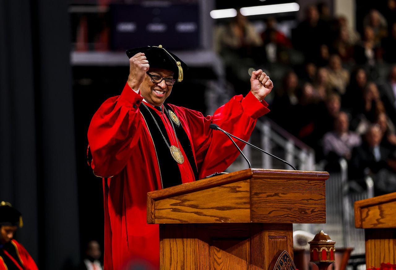 Neville Pinto in a cap and gown raises his hands over his head in triumph from the dais.