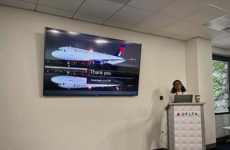Prissha Chawla stands behind a podium with a presentation screen behind her 