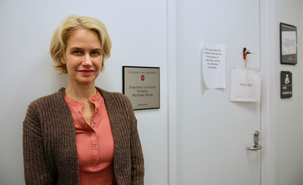 Head shot of Thea Langsam standing next to the entry door to a library nursing room and a wall plaque with the words, "University of Cincinnati, Dorthea Langsam Nursing Mothers' Room, dedicated 2023."