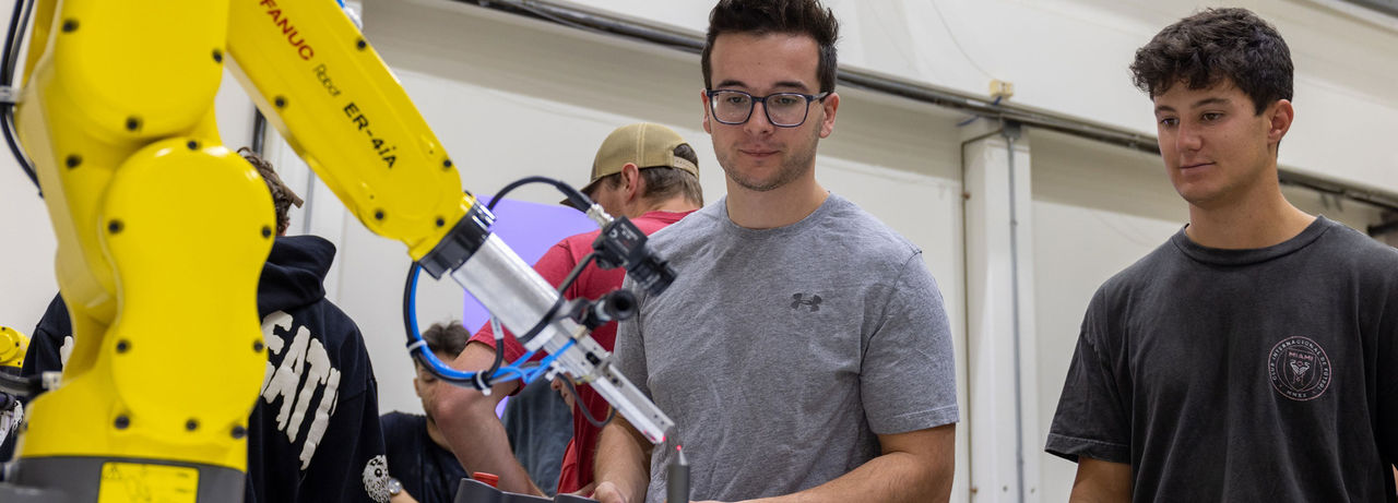 Two individuals observing a robotic arm in a workshop setting, with focus and interest.