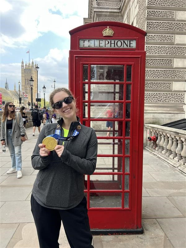 UC Alum Morgan Passek stands in front of a red phonebooth holding a gold medal