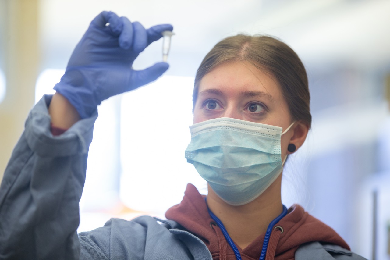 A student in a facemask and gloves holds up a vial in a lab.