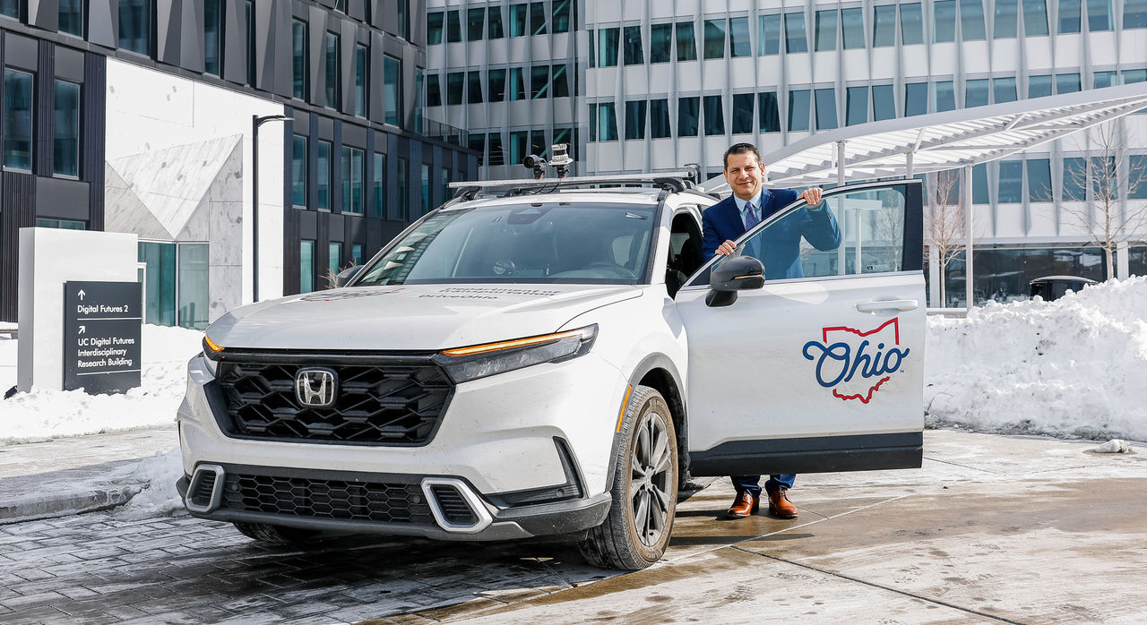 Munir Nazzal stands at the door of a Honda vehicle with the Ohio insignia in front of UC's Digial Futures. 