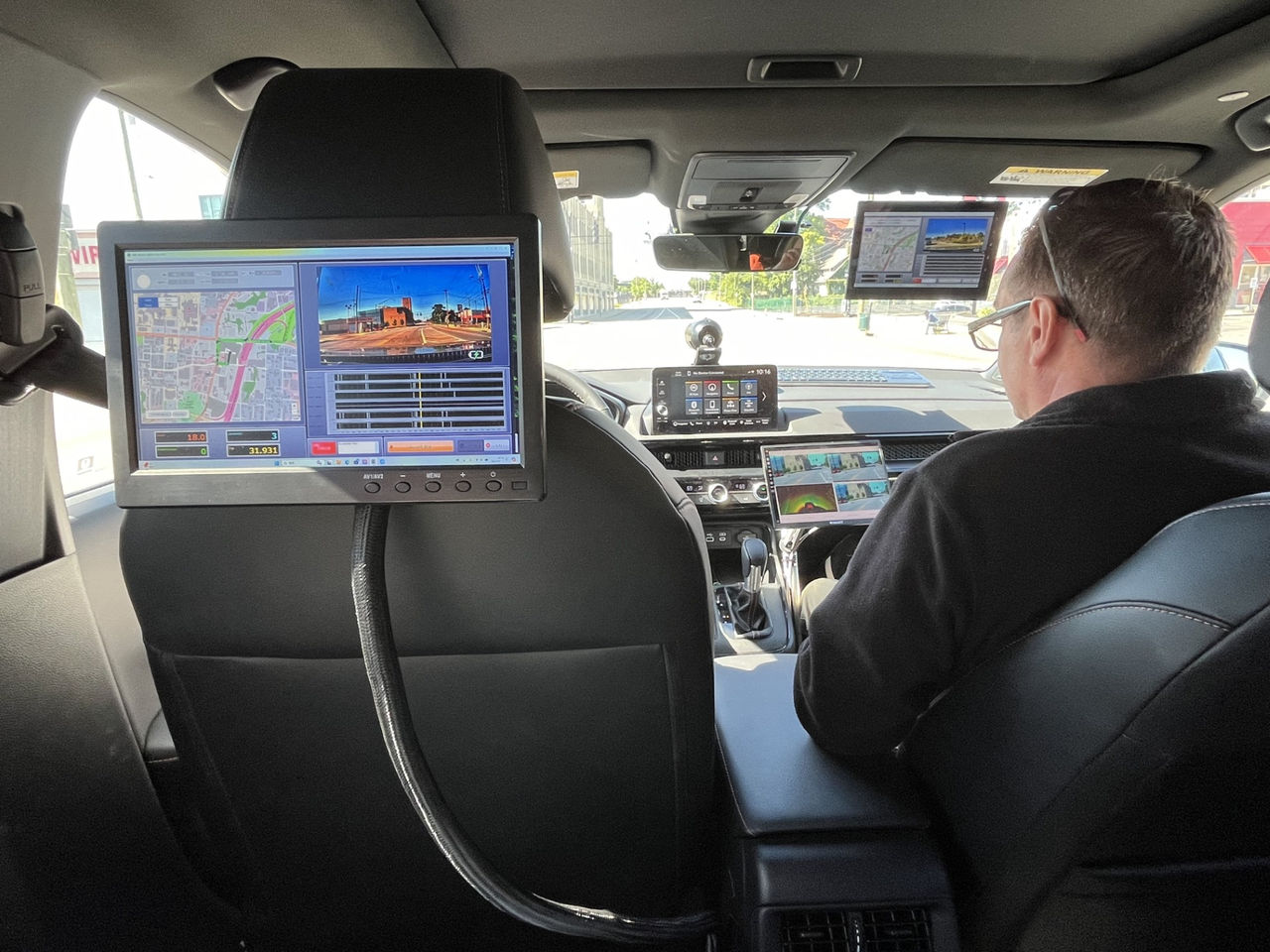 A backseat view of a car with computer screens shows cameras and GPS maps collecting road data.