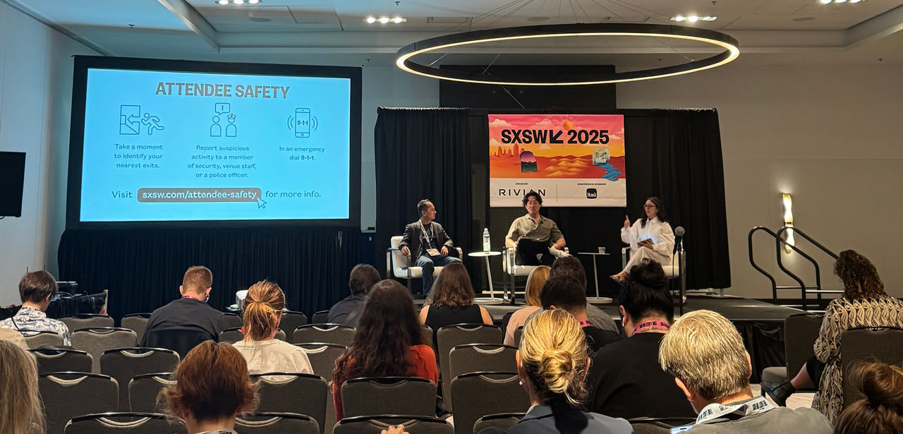 Three people are seated on stage in front of a banner that reads "SXSW 2025." The banner features abstract desert and sunset imagery and logos for Rivian and Itaú as sponsors. The panelists are sitting in white chairs, with one person gesturing while speaking, and the other two listening. The audience is partially visible in the foreground. The background consists of black curtains and a partial view of a presentation screen on the left. 