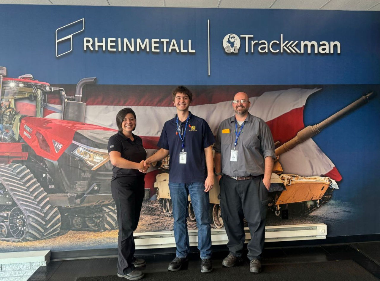 william walker stands in the center with two co-workers from American Rheinmetall standing on either side of him. Behind all of them is ap hoto of a tank and agriculatural equipment and the company logo up top