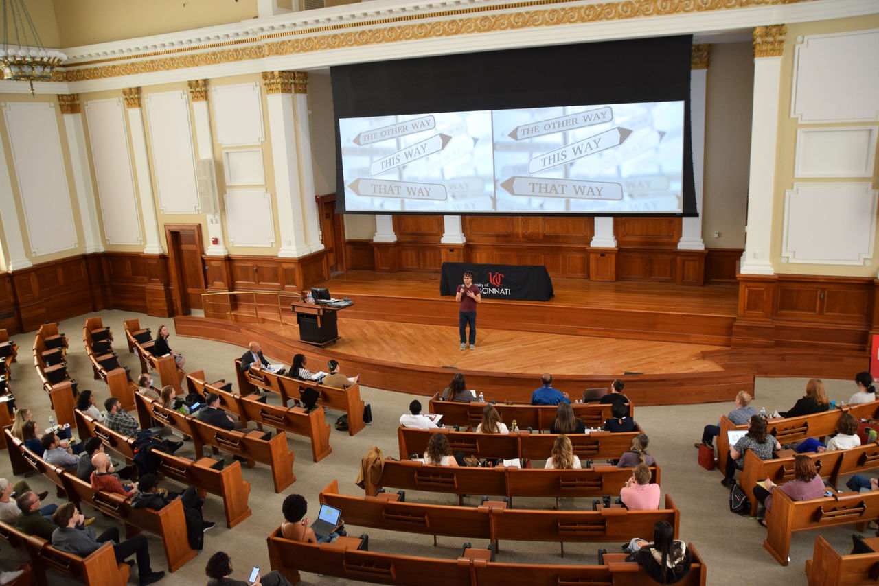 large auditorium with many people in circular style seating watching a speaker take centerstage