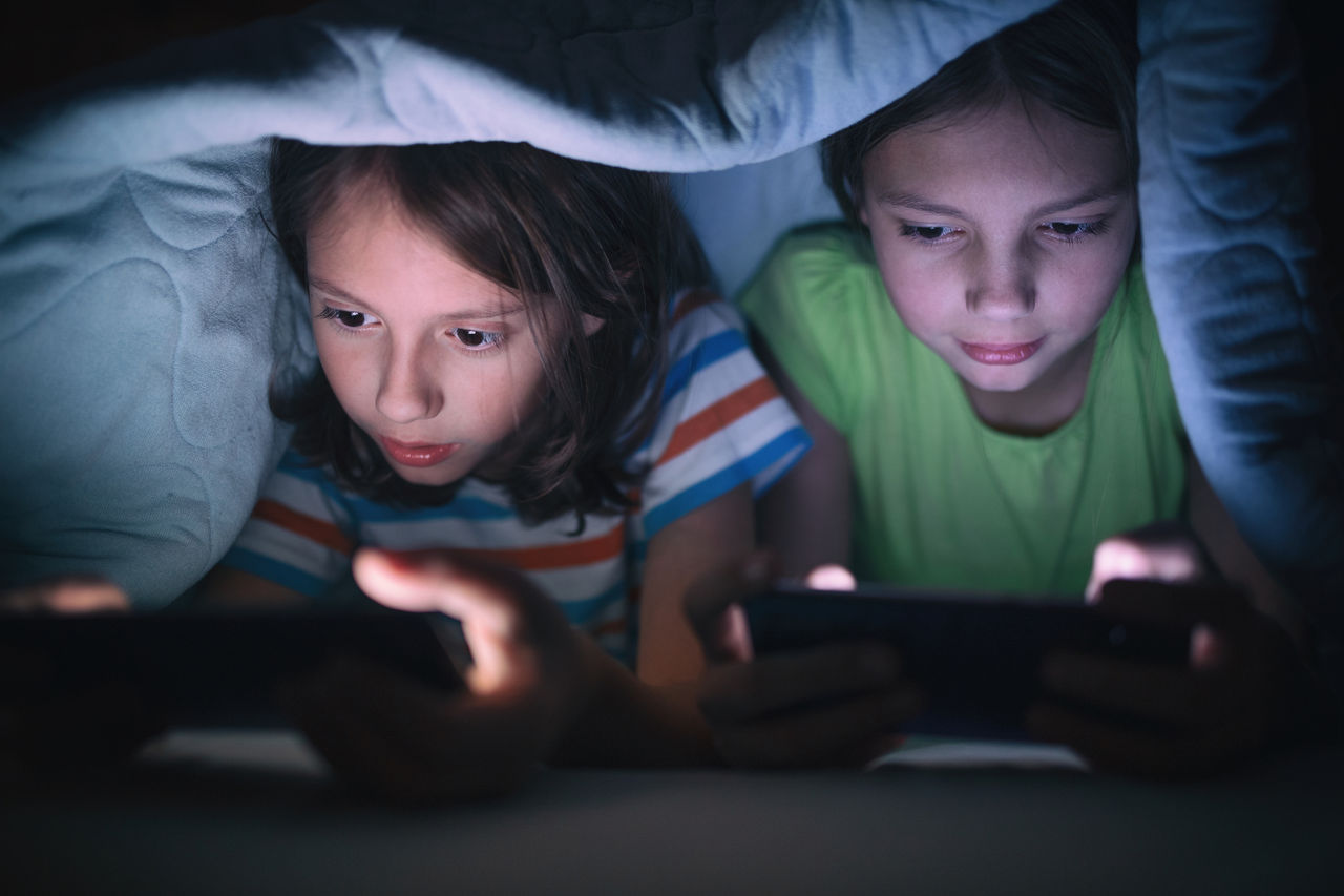 Two kids online gaming on devices while under a blanket 