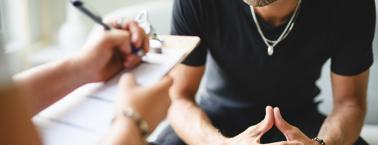 A woman with a clipboard takes notes in front of a man with his hands clasped anxiously in front of him.