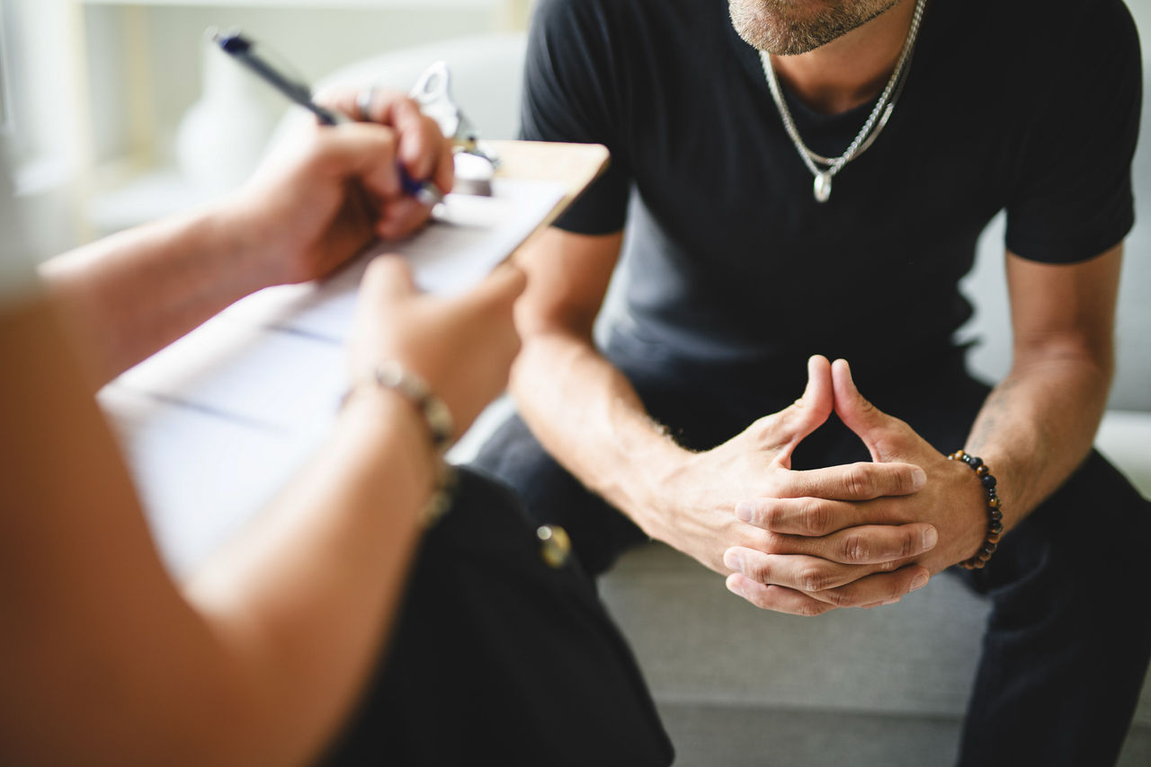 A woman with a clipboard takes notes in front of a man with his hands clasped anxiously in front of him.