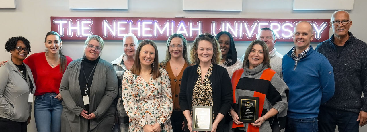 A group of twelve adults stand closely together indoors in front of a neon sign that reads ‘THE NEHEMIAH UNIVERSITY.’ Two people in the front row hold black and gold plaques, suggesting an awards or recognition event. The group is dressed in business‑casual clothing in a variety of colors and patterns.