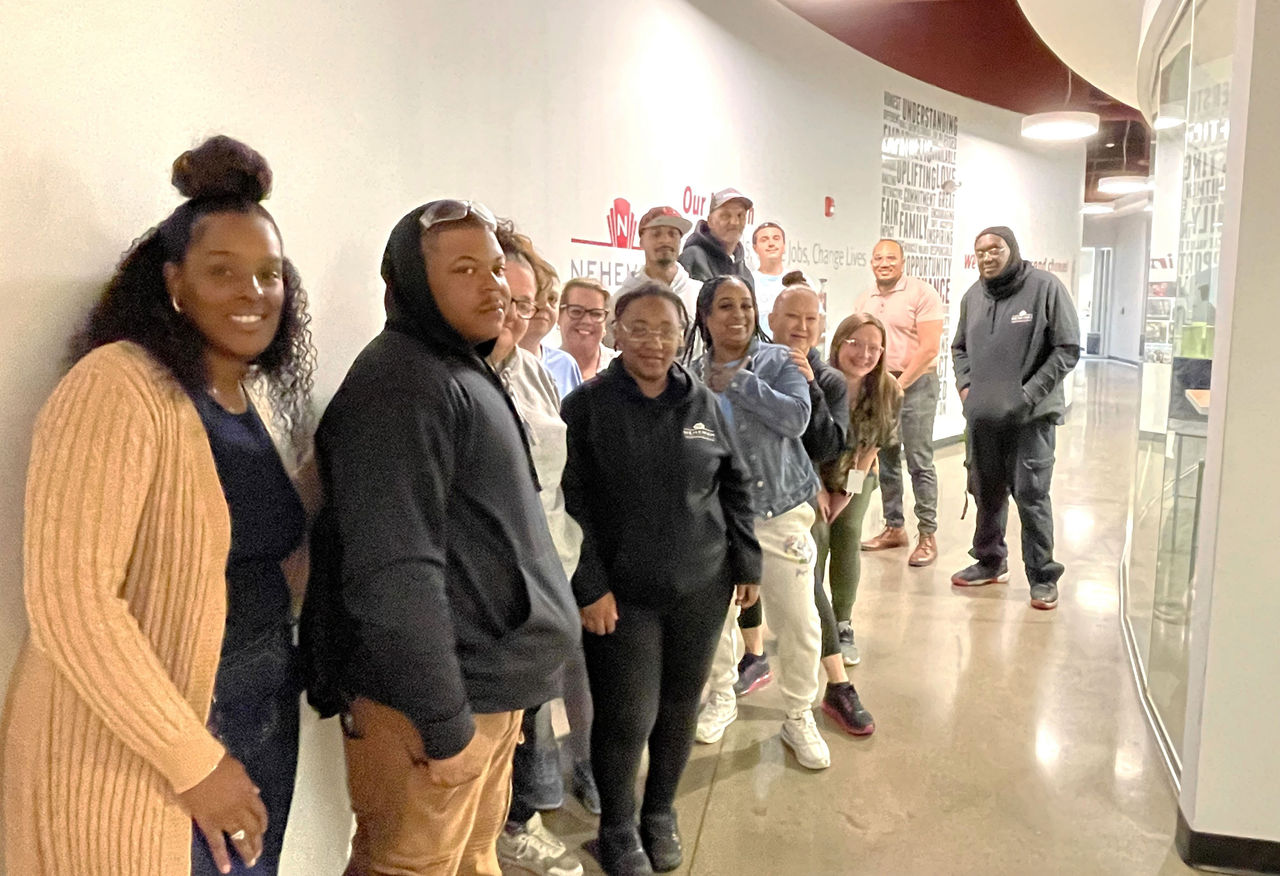 A diverse group of people stands in a line along a bright hallway, posing for a casual group photo. Their faces are blurred for privacy. Some wear hoodies and casual pants, while others are in sweaters and jeans. The polished floor reflects the overhead lights, and text and graphics are visible on the wall behind them.