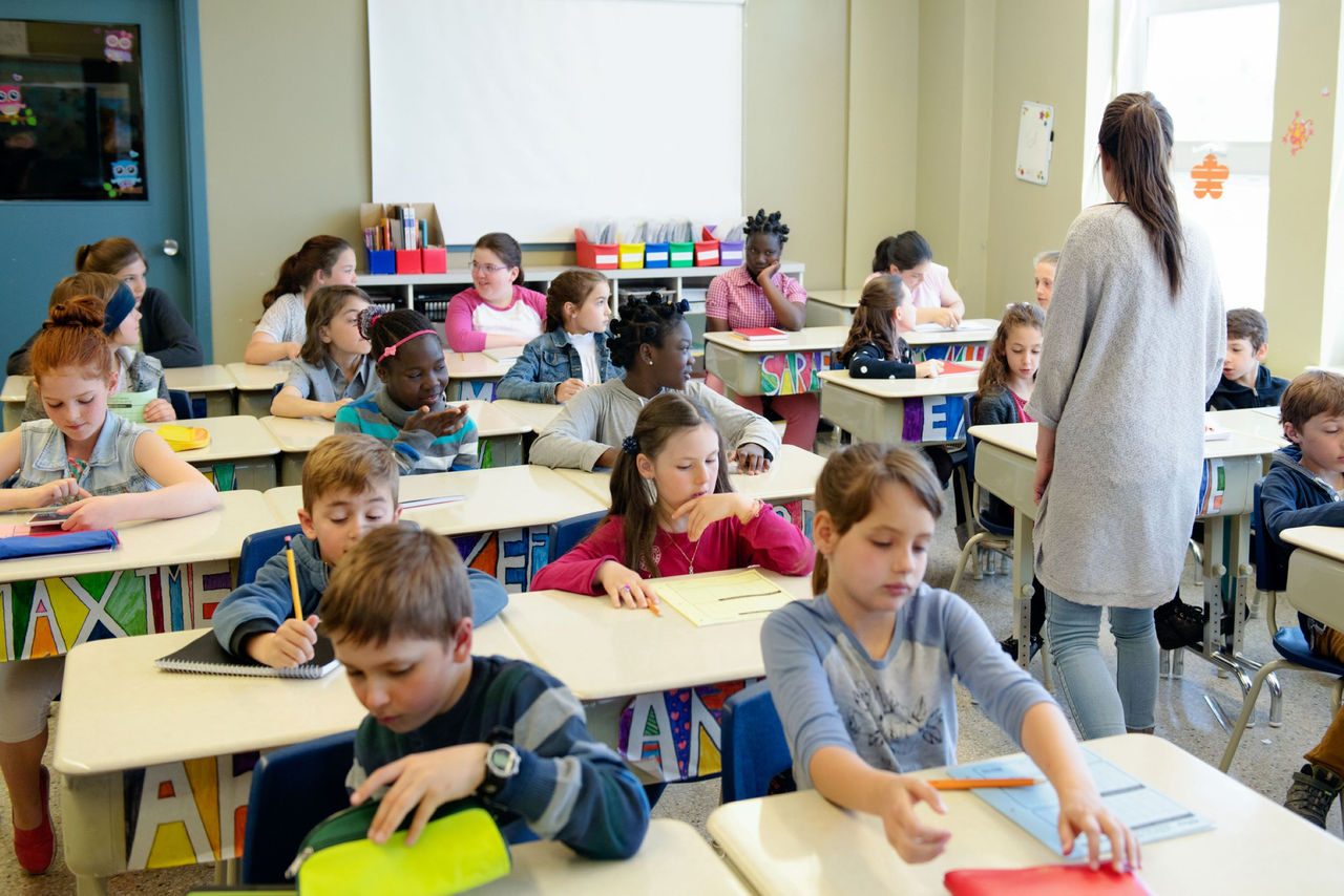 About two dozen elementary students sitting at classroom desks as their femaile teacher walks among them.