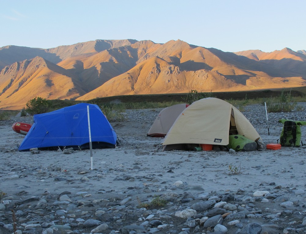 Tents are surrounded by an electric fence on a sandbar with mountains in the background.