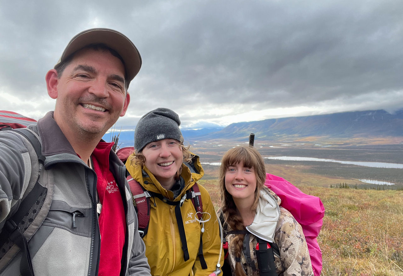 Three people in backpacks and rain jackets pose for a selfie on the Arctic landscape with mountains in the backround.