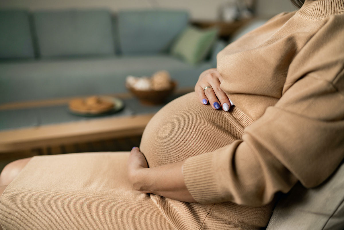 A pregnant woman wearing a tan sweater sits in a chair and rests her hands on her belly
