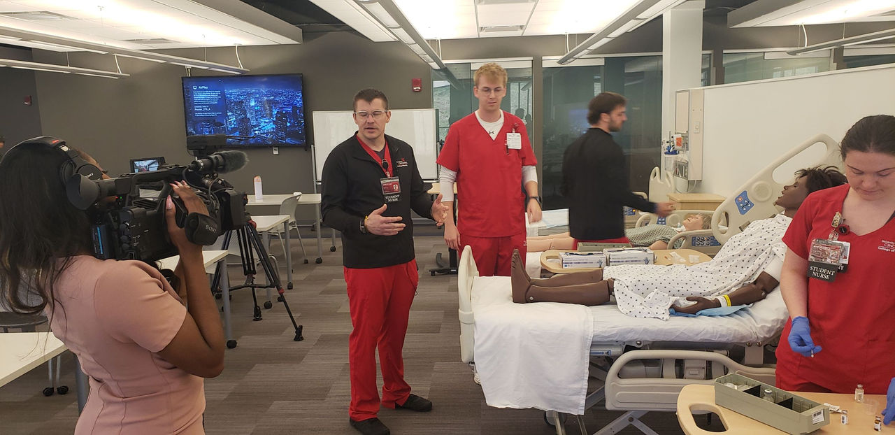 Reporter hold video camera and aims it at nursing student for an interview. Three other nursing students seen in the background working with a simulated patient.