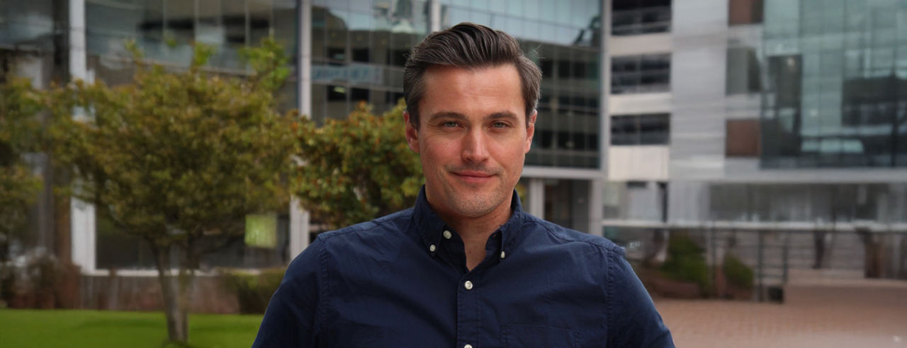 John Kelly stands outside with glass buildings and trees behinds him