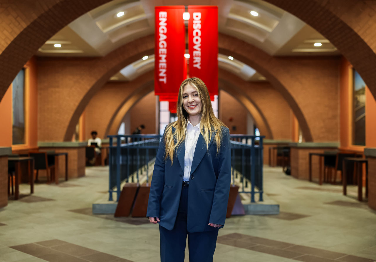 Savannah Dickens dressed in a blue paint suit standing in the lobby of Mantei Center on the UC campus. Banners with the words Discovery and Innovation hang in the background.  