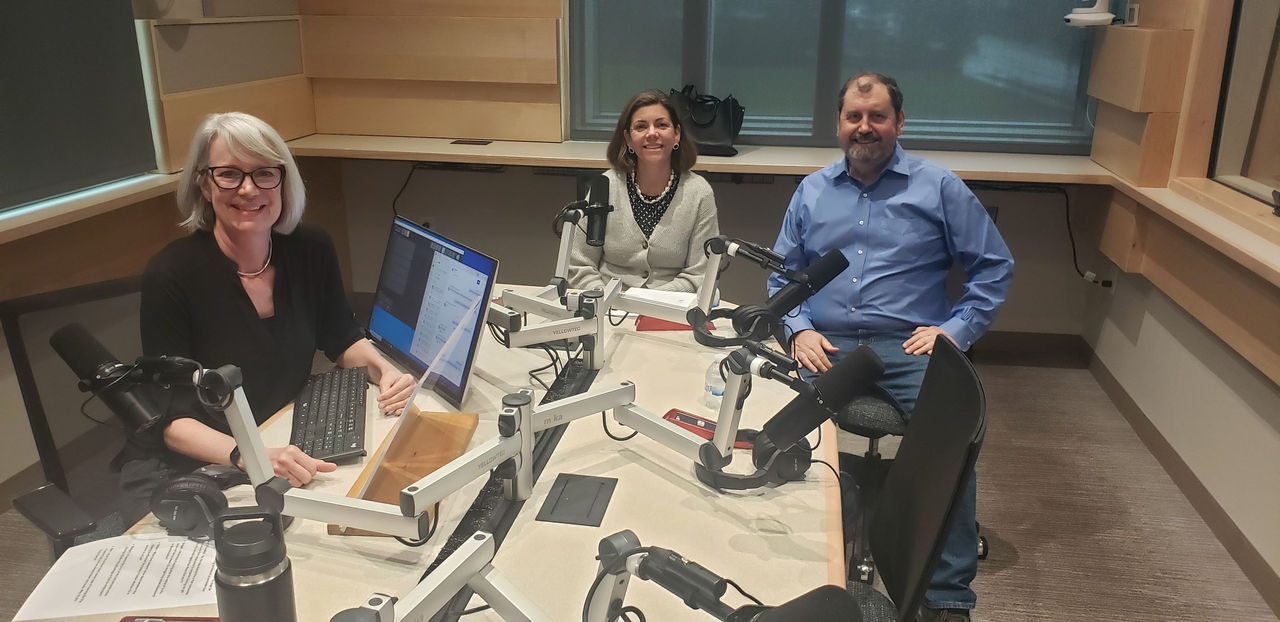 Journalist Lucy May is seated behind a computer with guests Bridget McGraw and Neil Taylor sitting nearby behind microphones