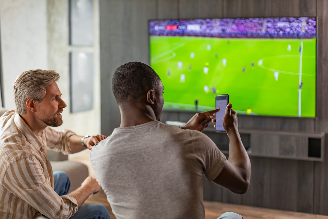 Two men watch a sports match on a big screen TV and one holds up a cell phone to place a sports bet.
