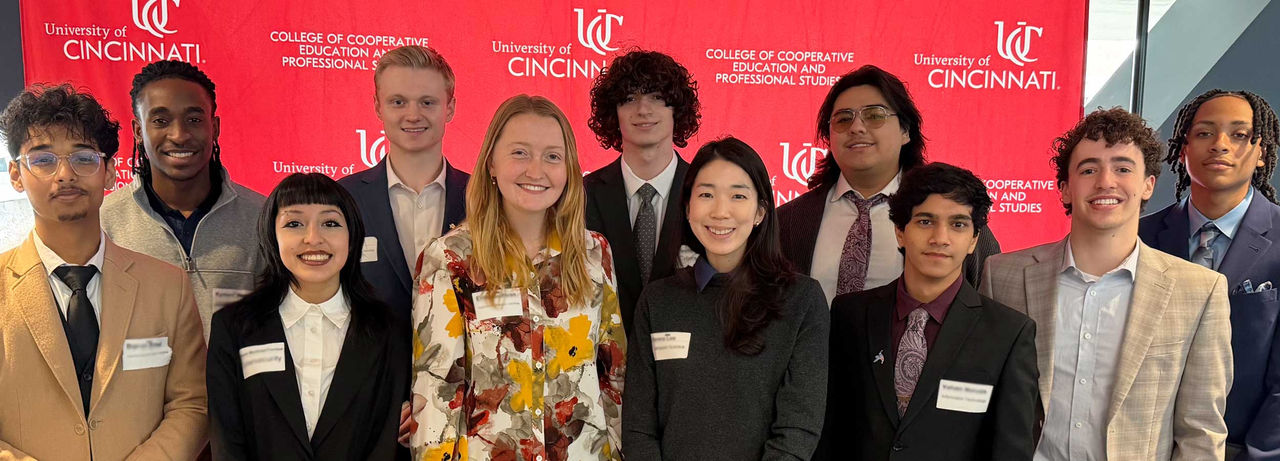 Group of eleven students wearing professional business attire stand in two rows in front of a red University of Cincinnati backdrop that reads “UC College of Cooperative Education and Professional Studies.” They are posed closely together, smiling, with name tags visible on their jackets and shirts.