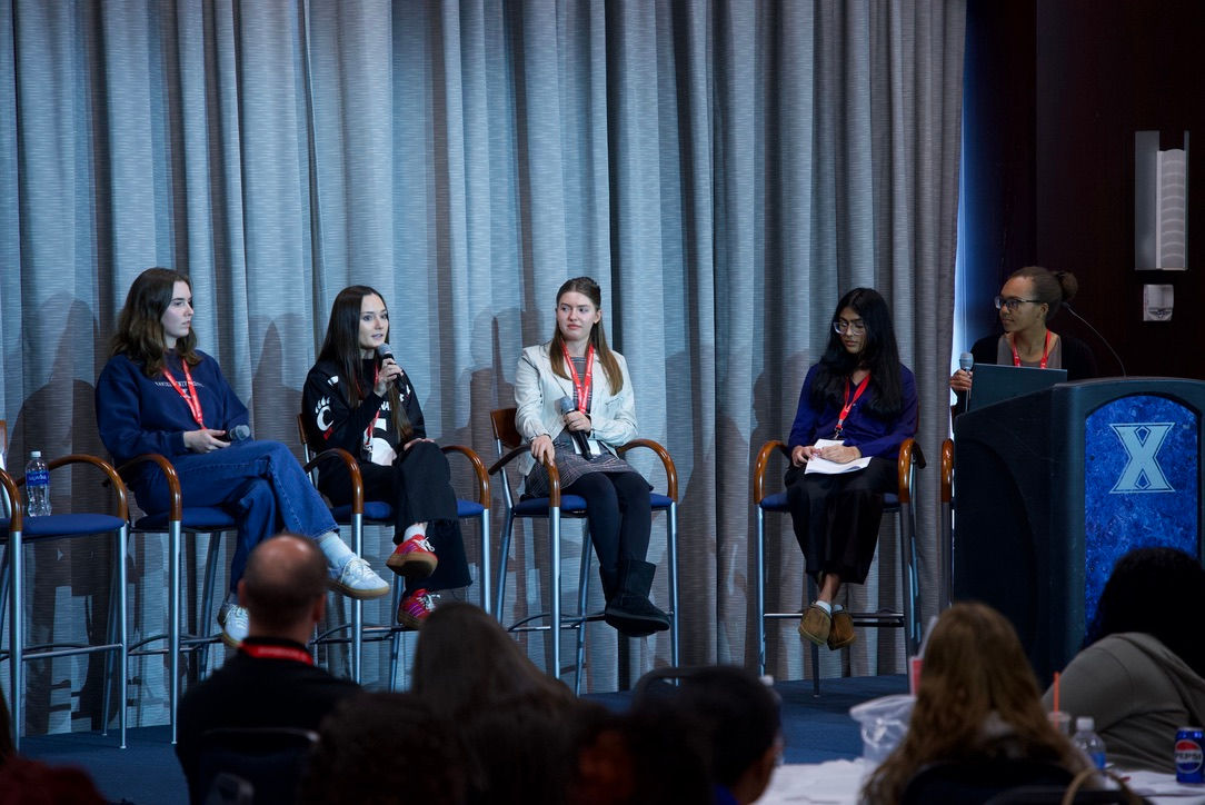 Hannah Laman is one of 5 women that sits on a stage in a panel, holding a microphone. 