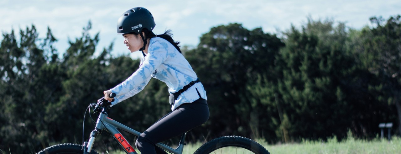 A woman wearing a black helmet rides a bike outside with trees and grass in the background behind her