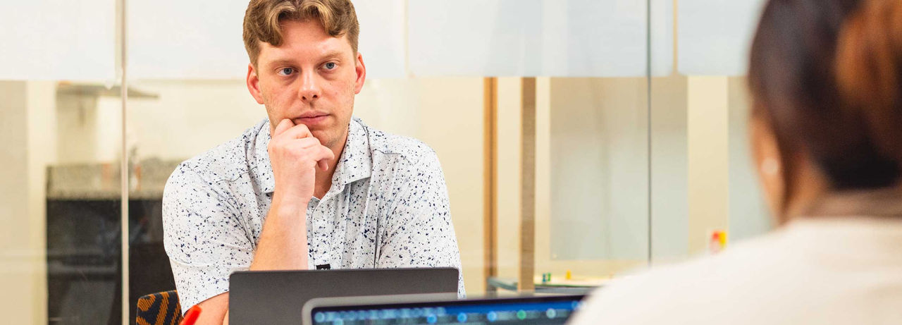 Evan Kamp sits at a table in a bright indoor workspace, resting their chin on their hand and looking toward another person in the foreground. Laptops are open in front of both individuals, suggesting a meeting or tutoring session in a modern office or study area.
