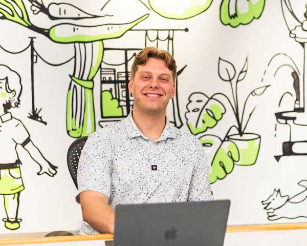 Evan Kamp sits at a desk working on an open laptop. Behind them is a bright white wall with bold green and black cartoon-style illustrations of people and plants.