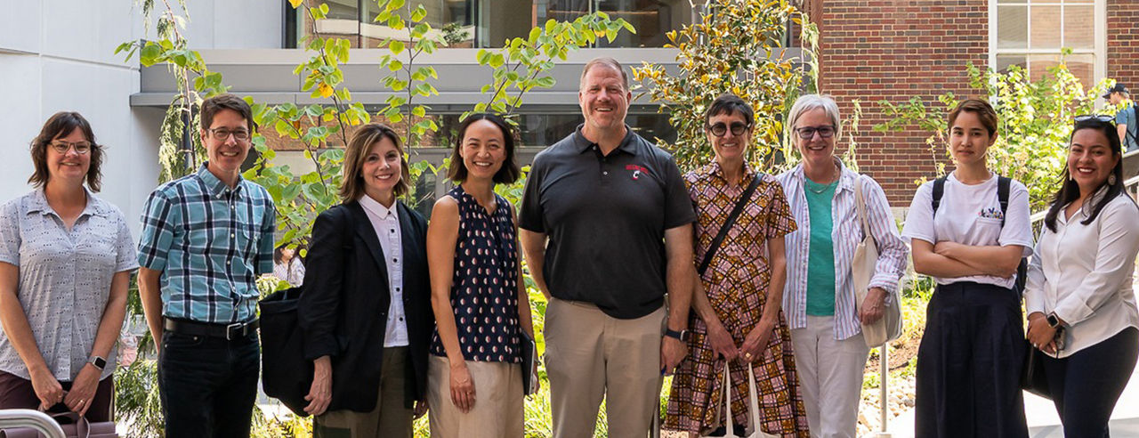 a group of people stand in a line in front of trees and a University of Cincinnati building