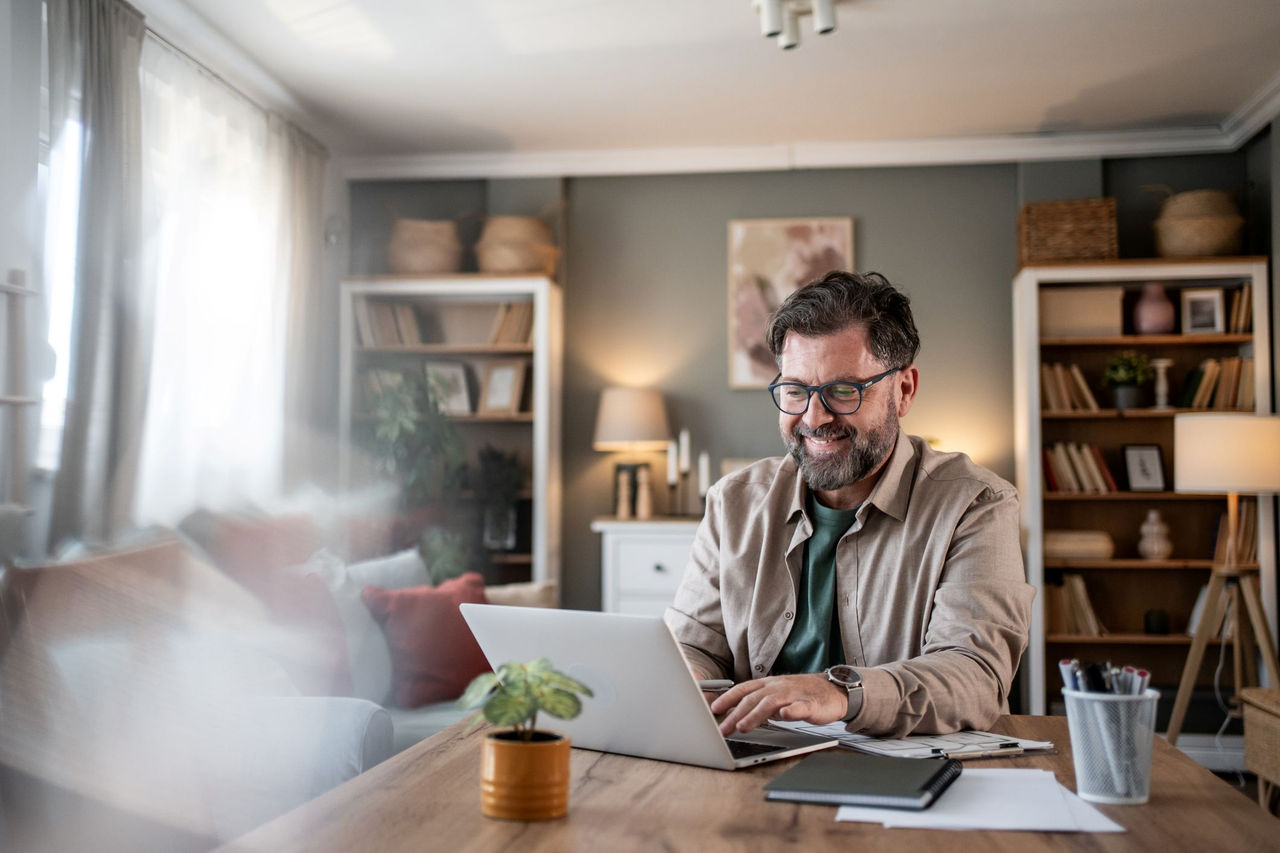 Man sitting at a table with a laptop working and smiling. He is in his home.