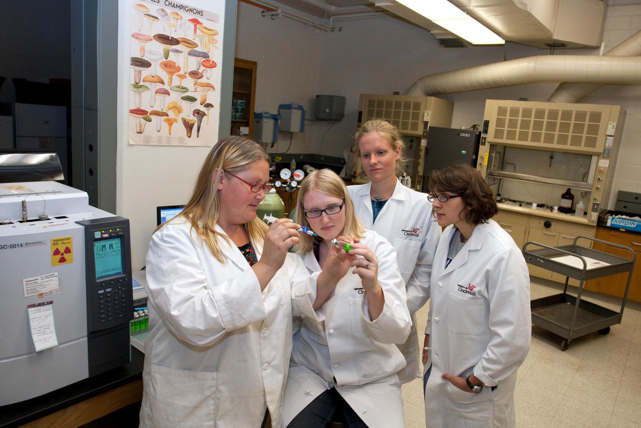 Amy Townsend-Small and students in white labcoats crowd around a sample in a lab.
