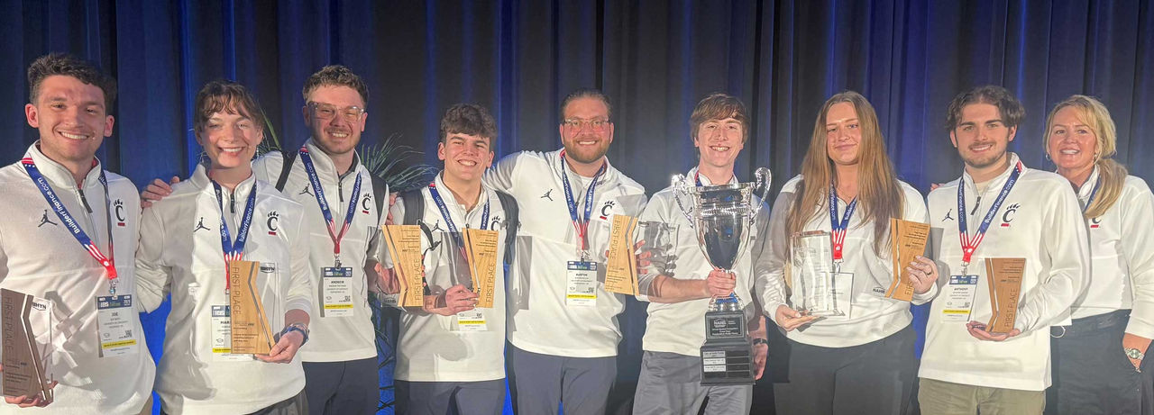 A large group of UC students in matching white quarter-zip shirts with the Bearcat logo stand shoulder to shoulder on a stage in front of dark curtains, each wearing conference lanyards and holding wooden “First Place” plaques, while two students in the center display oversized championship trophies.